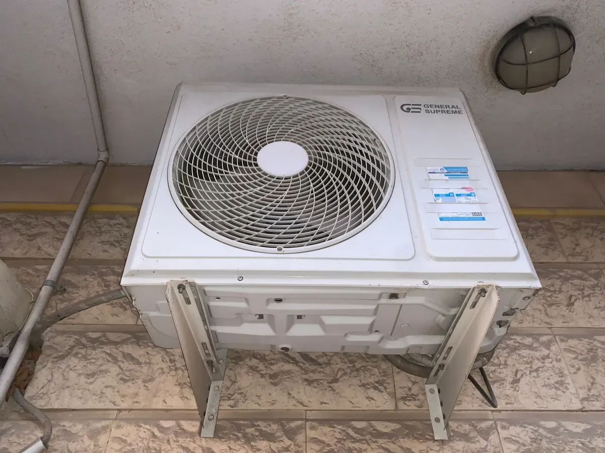 وحدة سبليت خارجية بيضاء مستعملة على حامل معدني فوق أرضية مبلطة.